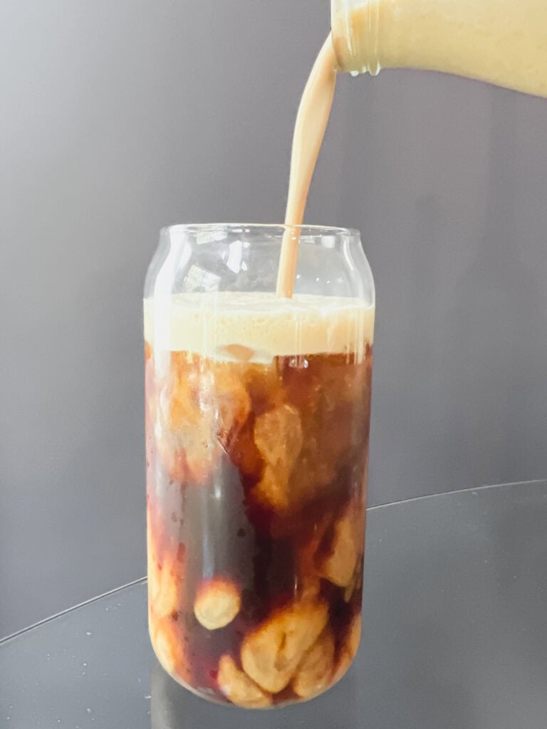 A clear glass of iced coffee topped with homemade salted pumpkin coffee creamer. The creamy foam swirls into the coffee, dusted with pumpkin pie spice and a pinch of flaky Redmond Real Salt, creating a cozy fall drink that looks rich, smooth, and inviting.