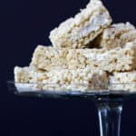 Rice Krispies treats cut into squares arranged on a glass cake stand with a fall linen napkin under it.