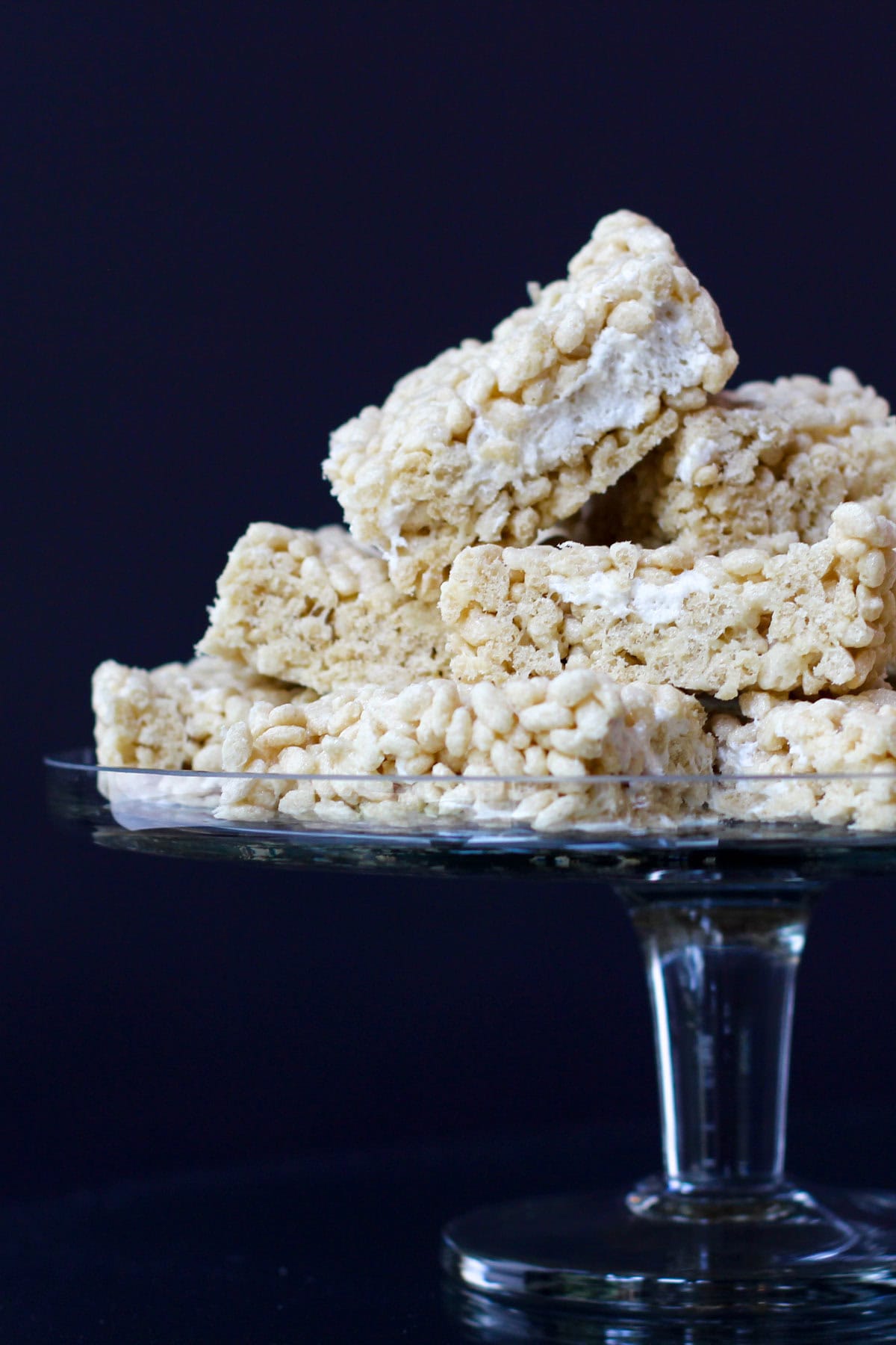 Rice Krispies treats cut into squares arranged on a glass cake stand with a fall linen napkin under it.