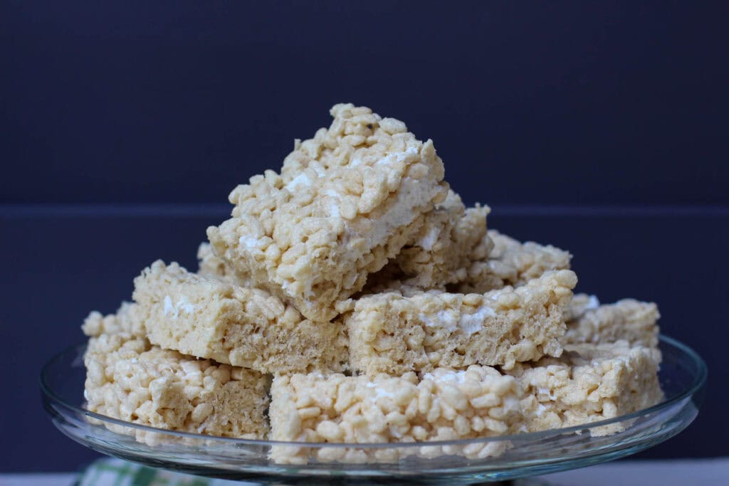 Rice Krispies treats cut into squares arranged on a glass cake stand with a fall linen napkin under it.