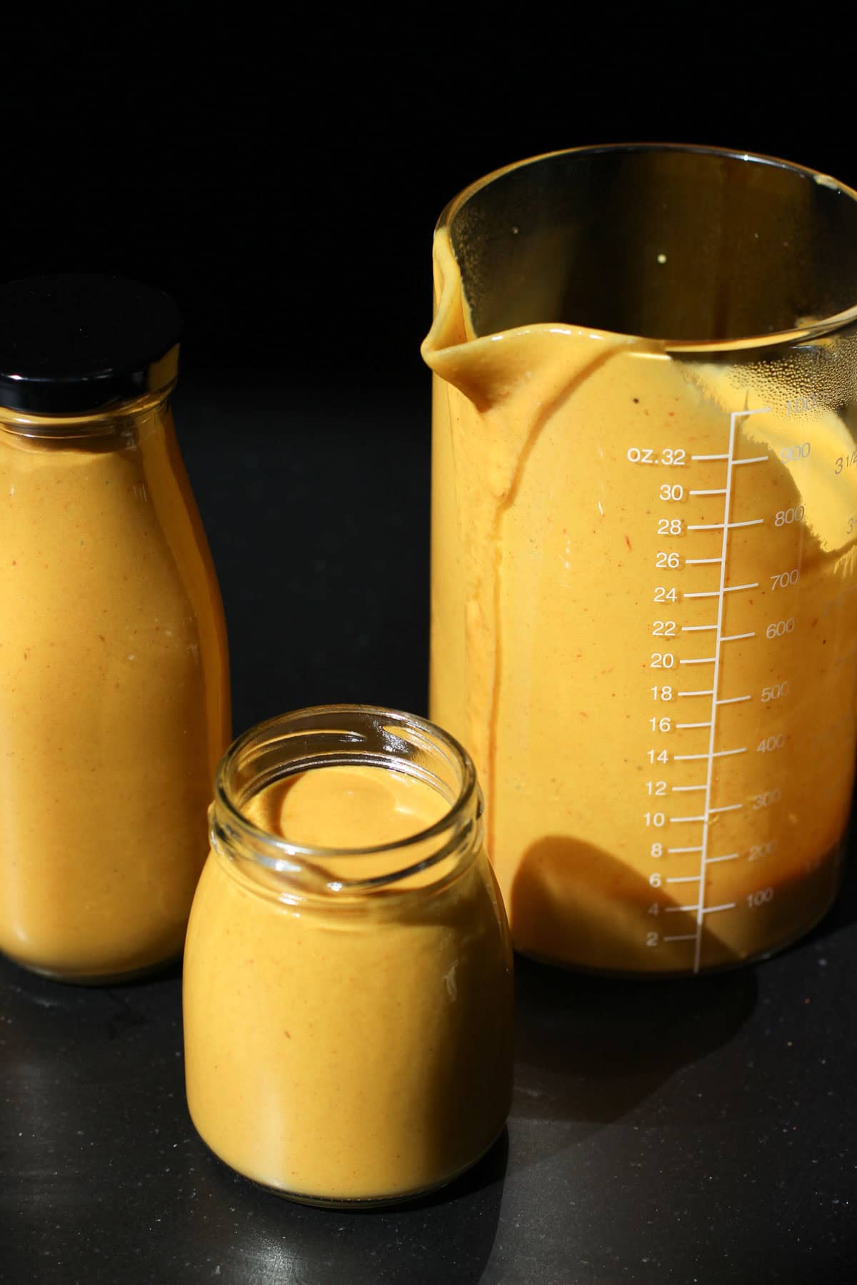 Creamy Carolina Gold BBQ Sauce is in three glass jars. Some sauce drips down the side of one jar. They are all on a black countertop.