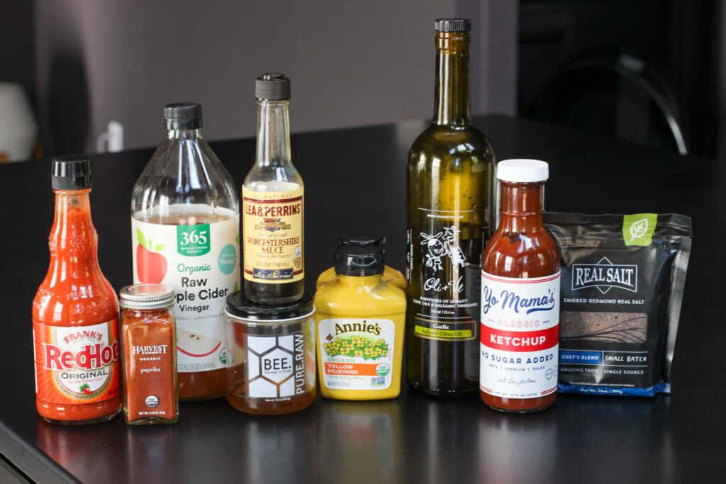 Tomato ketchup, mustard, apple cider vinegar, hot sauce, soy sauce, smoked salt, olive oil, and garlic oil on black table.