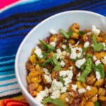 A white bowl filled with creamy elote corn topped with Cotija cheese, cilantro, and chili powder on a colorful woven cloth, ready to serve for entertaining.