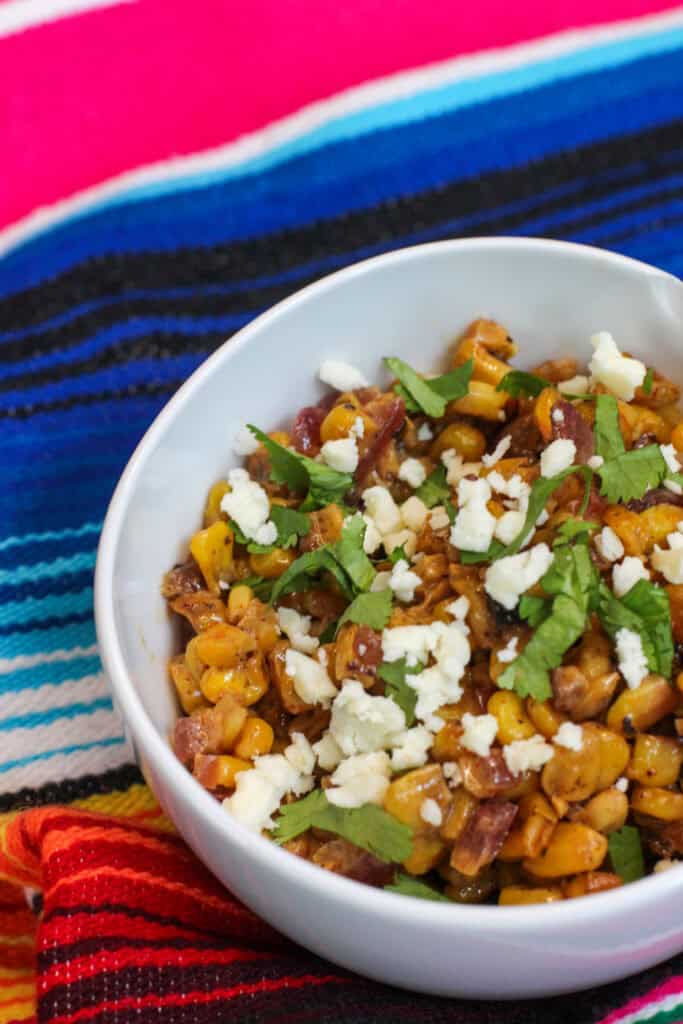 A white bowl filled with creamy elote corn topped with Cotija cheese, cilantro, and chili powder on a colorful woven cloth, ready to serve for entertaining.