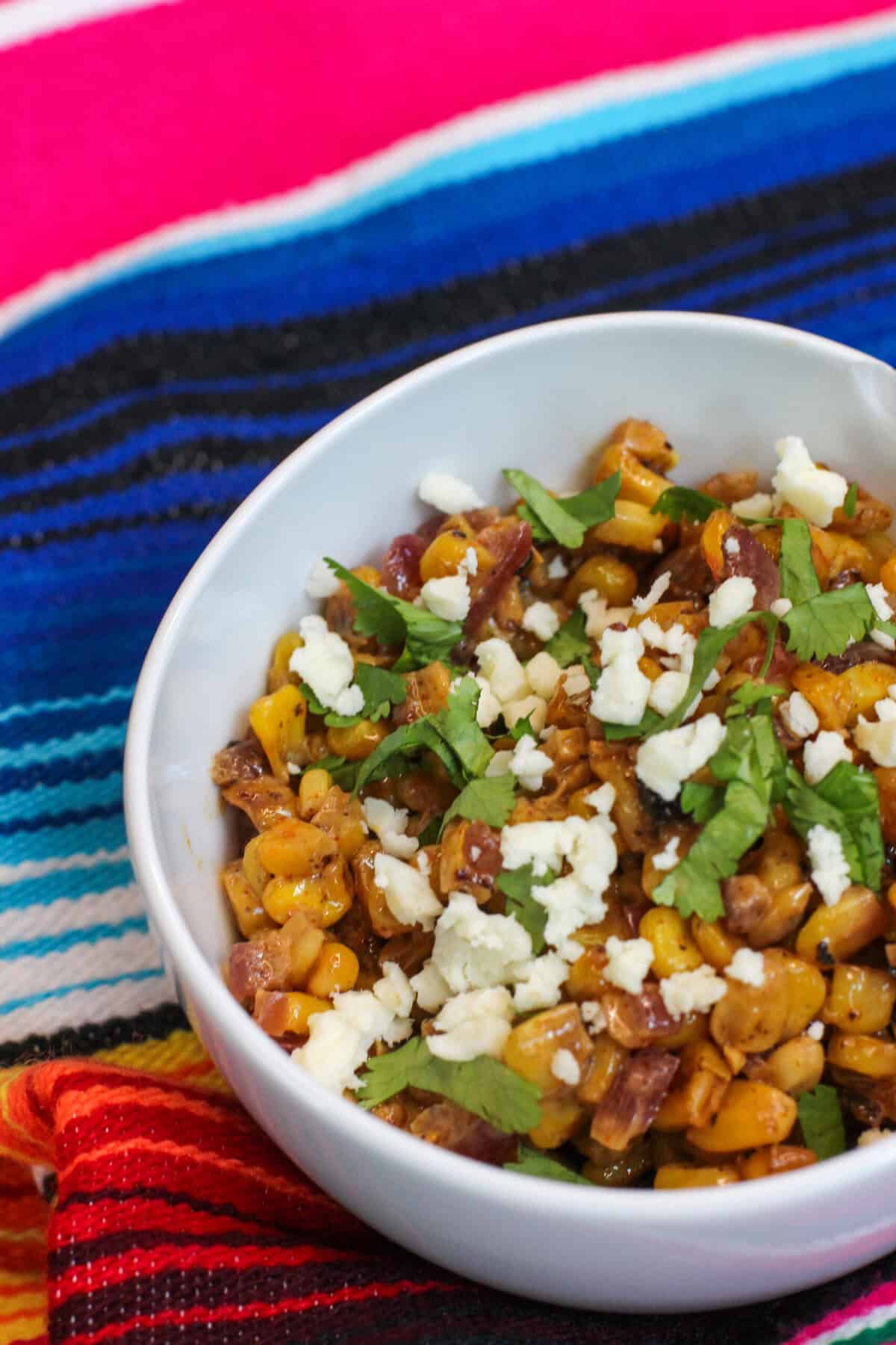 A white bowl filled with creamy elote corn topped with Cotija cheese, cilantro, and chili powder on a colorful woven cloth, ready to serve for entertaining.