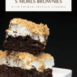 Two stacked s’mores brownies topped with toasted marshmallow and graham cracker crumble, showing layers of fudgy chocolate brownie, gooey marshmallow, and crunchy crumbs against a dark background.