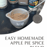 Overhead photo of a small gray measuring cup filled with homemade apple pie spice on a black surface, surrounded by open spice jars, a whisk, and metal measuring spoons. A gold lid labeled “Apple Pie Spice” rests nearby.