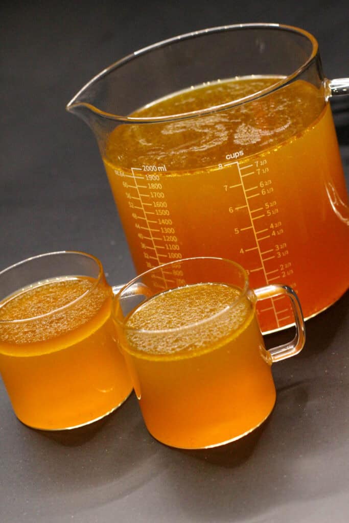 A glass measuring cup filled with golden homemade bone broth next to smaller glass cups for sipping, set on a dark countertop.