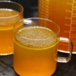 A glass measuring cup filled with golden homemade bone broth next to smaller glass cups for sipping, set on a dark countertop.