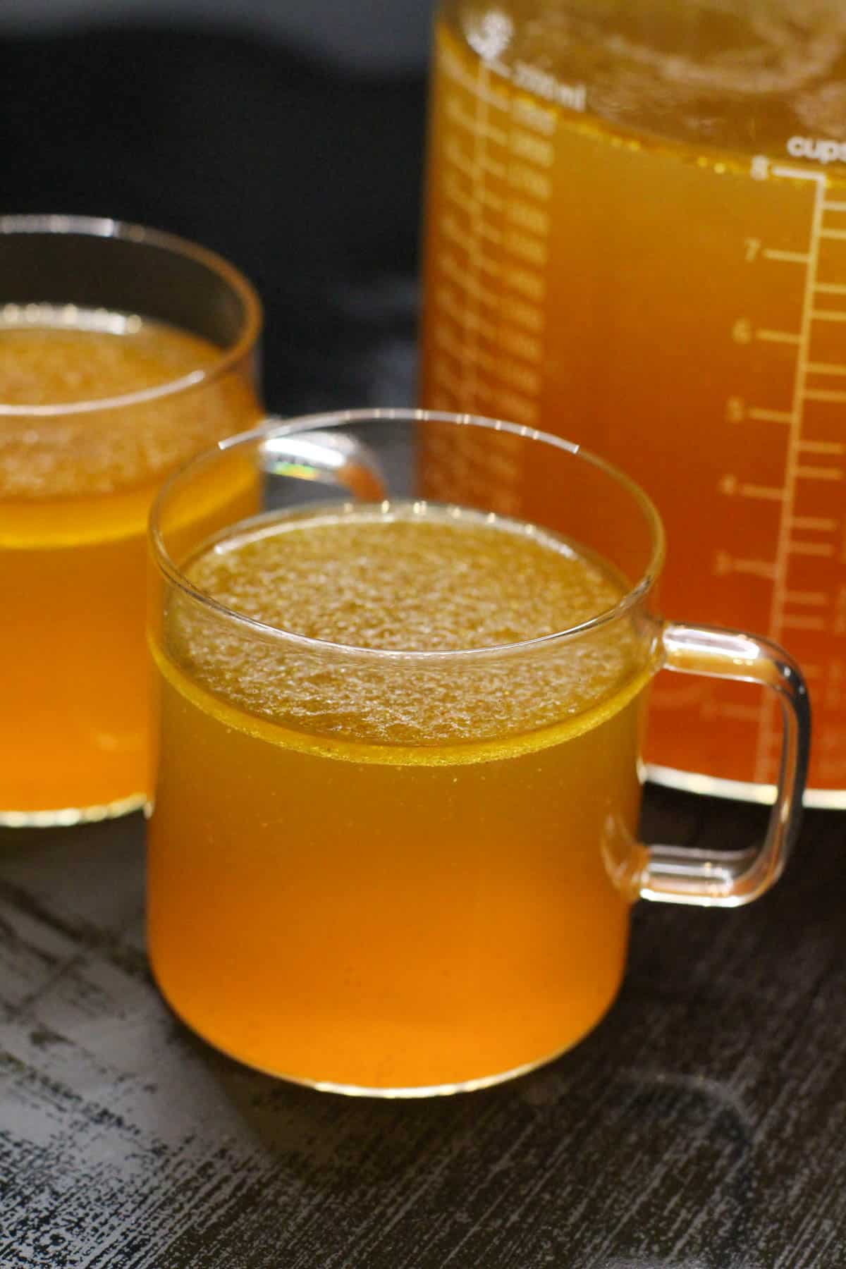 A glass measuring cup filled with golden homemade bone broth next to smaller glass cups for sipping, set on a dark countertop.