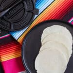 Freshly pressed homemade masa tortillas on a black plate beside a traditional cast-iron tortilla press and colorful striped cloth, ready for cooking or serving.