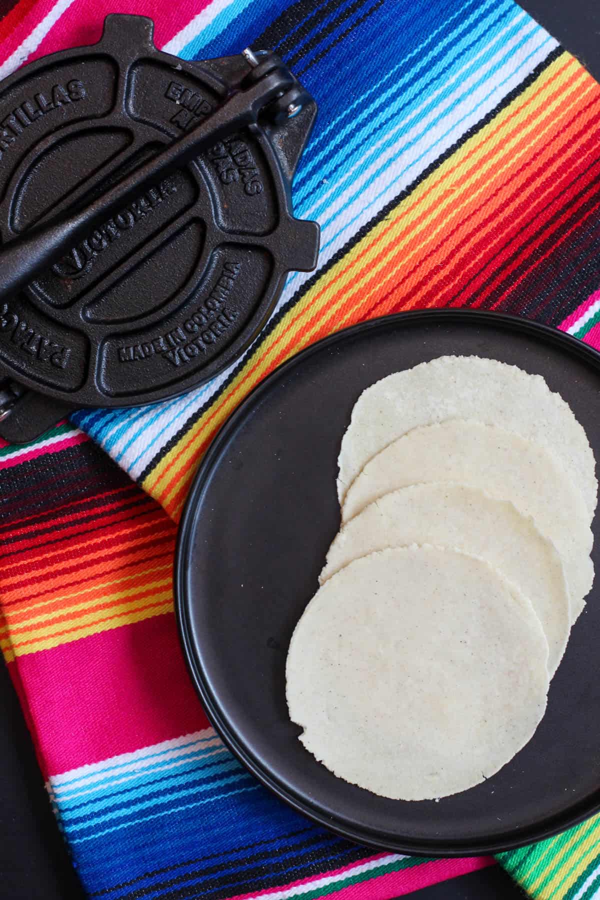 Freshly pressed homemade masa tortillas on a black plate beside a traditional cast-iron tortilla press and colorful striped cloth, ready for cooking or serving.