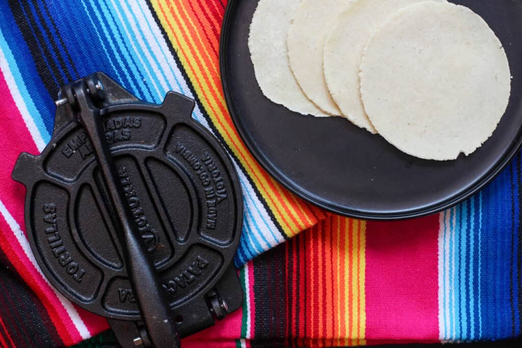 Freshly pressed homemade masa tortillas on a black plate beside a traditional cast-iron tortilla press and colorful striped cloth, ready for cooking or serving.