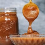 A spoon drizzling thick homemade apple butter into a glass bowl beside of mason jar of apple butter, set against a gray marble background.