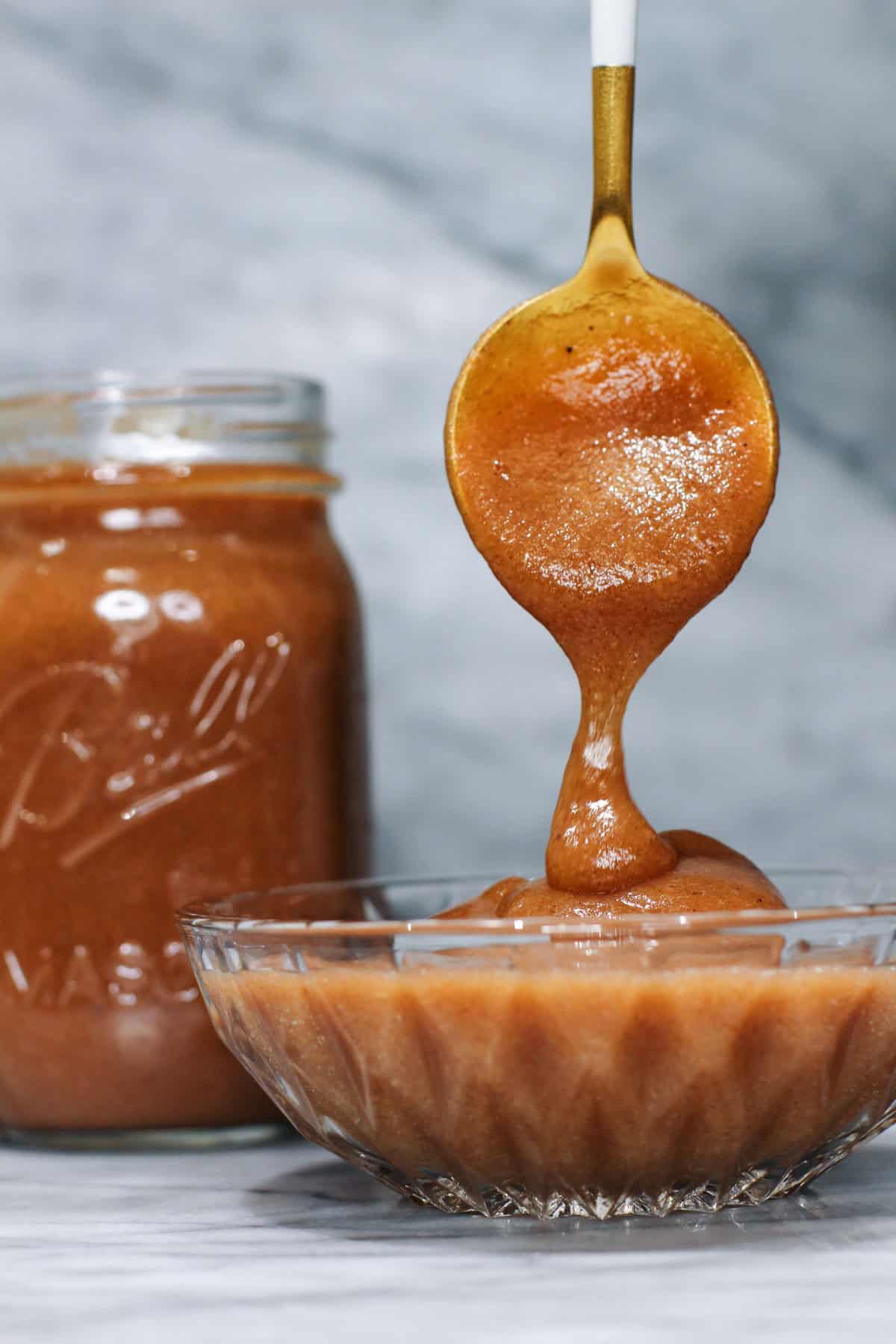 A spoon drizzling thick homemade apple butter into a glass bowl beside of mason jar of apple butter, set against a gray marble background.