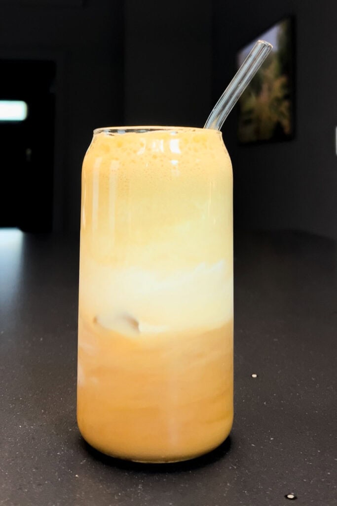 A glass of maple cinnamon shaken espresso with frothy layers and a glass straw, sitting on a dark countertop in natural light.