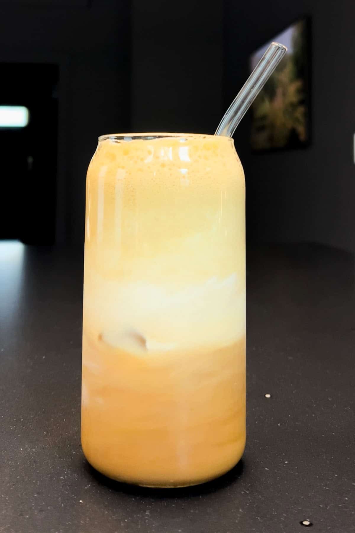 A glass of maple cinnamon shaken espresso with frothy layers and a glass straw, sitting on a dark countertop in natural light.
