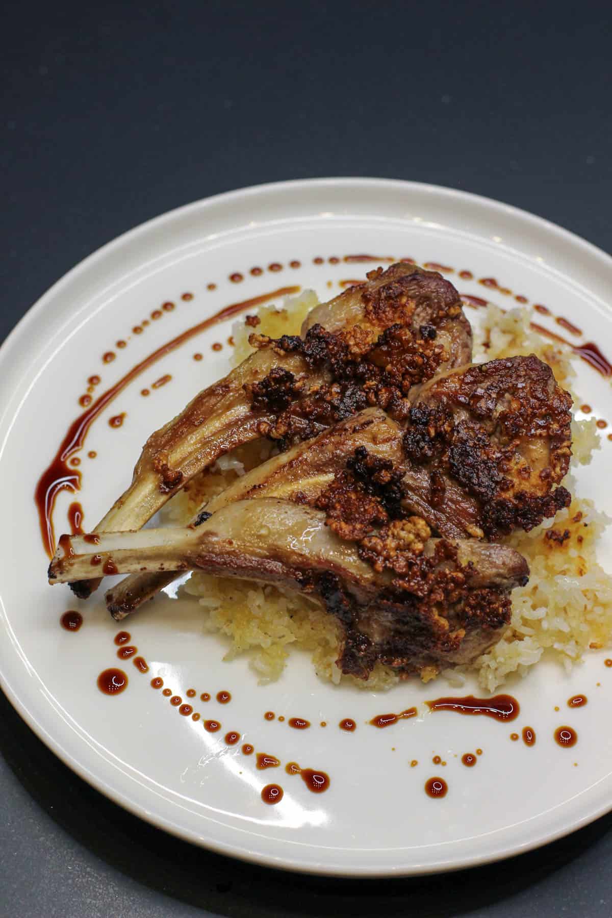 Rosemary Dijon crusted lamb lollipops served over a bed of fluffy rice and drizzled with balsamic reduction on a white plate.