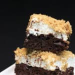 A stack of two s'mores brownies, topped with toasted marshmallows and a graham cracker crumble on a white plate sitting on a black countertop.