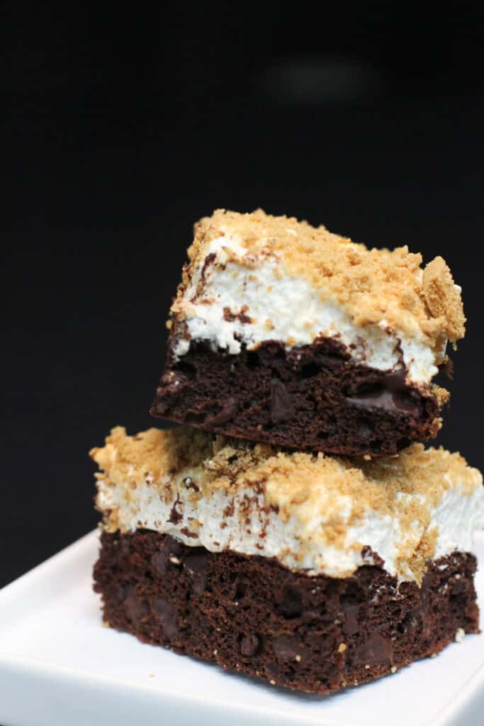 A stack of two s'mores brownies, topped with toasted marshmallows and a graham cracker crumble on a white plate sitting on a black countertop.