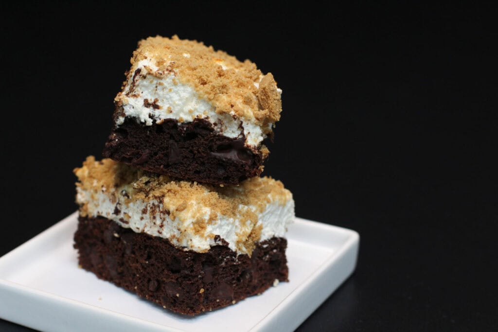 A stack of two s'mores brownies, topped with toasted marshmallows and a graham cracker crumble on a white plate sitting on a black countertop.