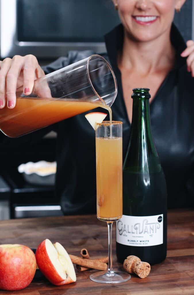 A woman pours homemade apple cider into a champagne flute beside apples, cinnamon sticks, and a bottle of sparkling wine on a wooden counter—perfect for entertaining.