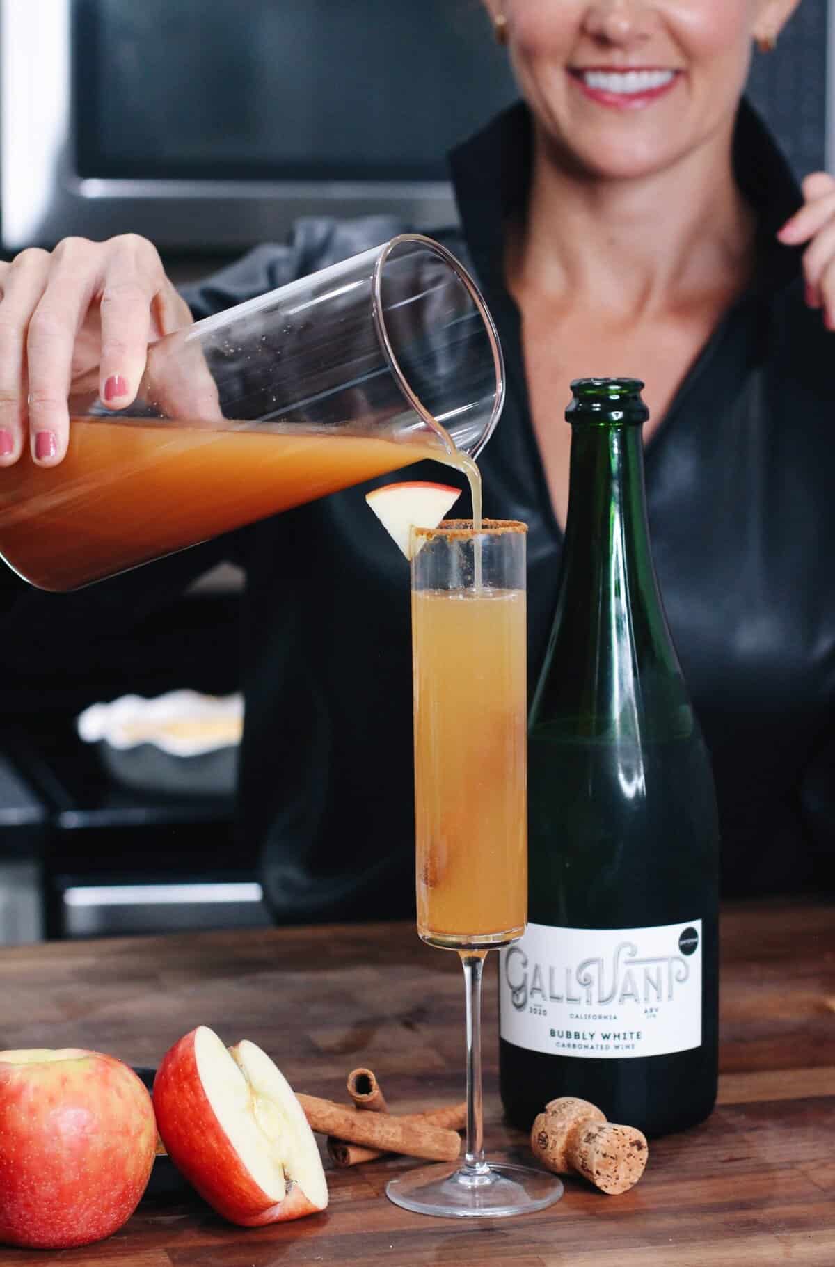 A woman pours homemade apple cider into a champagne flute beside apples, cinnamon sticks, and a bottle of sparkling wine on a wooden counter—perfect for entertaining.