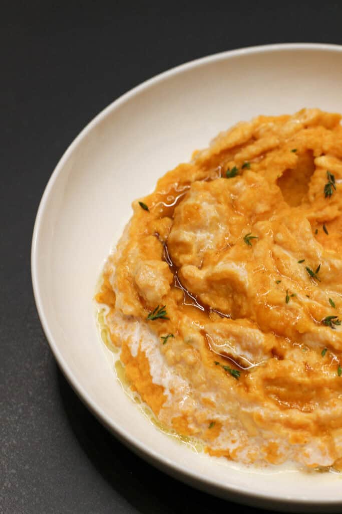 A close-up of creamy sweet potato mash swirled with whipped ricotta and drizzled with maple syrup, topped with fresh thyme leaves for an elegant, cozy presentation.