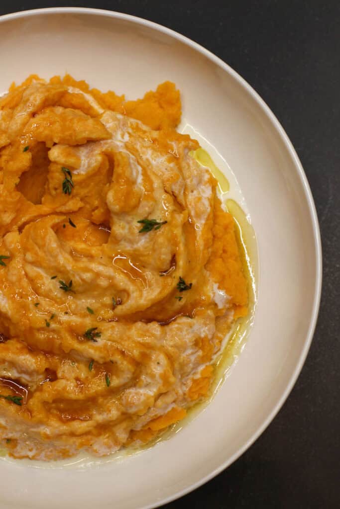 A close-up of creamy sweet potato mash swirled with whipped ricotta and drizzled with maple syrup, topped with fresh thyme leaves for an elegant, cozy presentation.