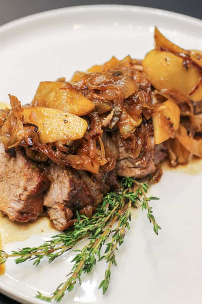 Slices of cider-braised pork loin topped with caramelized apple-onion compote and fresh thyme on a white serving platter.