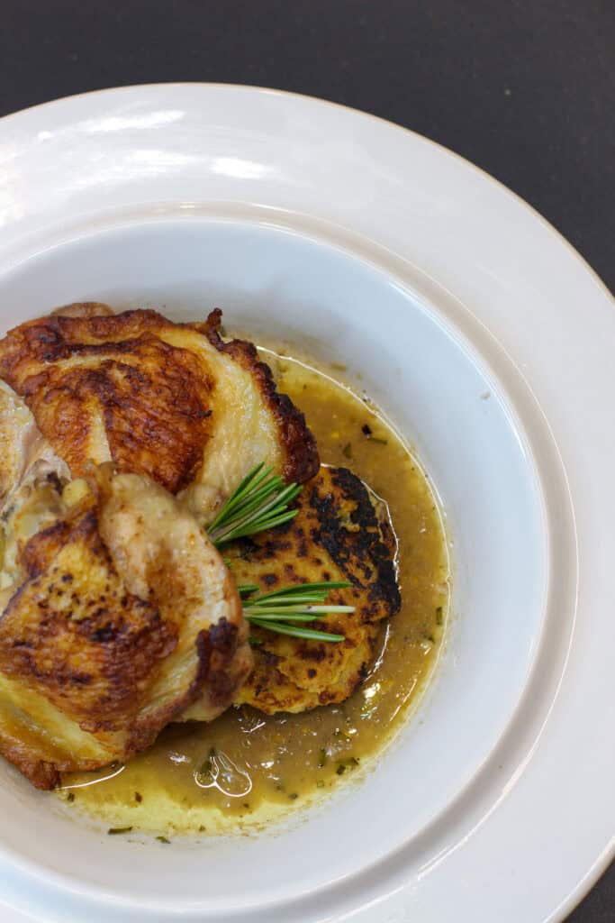 Plate of crispy-skinned chicken thighs served with rosemary jus and pan-seared polenta cakes on a white dish.