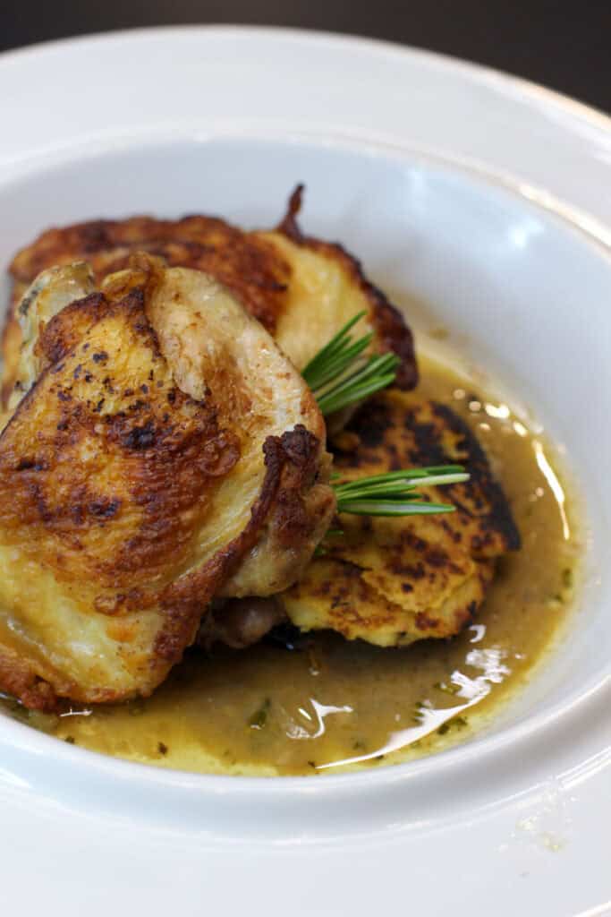 Plate of crispy-skinned chicken thighs served with rosemary jus and pan-seared polenta cakes on a white dish.