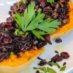 Roasted honeynut squash halves filled with wild rice, mushrooms, cranberries, and toasted pecans, garnished with fresh parsley on a white plate.
