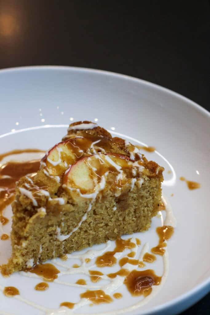 Close-up image of a slice of rustic apple and cornmeal cake drizzled with cider caramel and crème fraîche on a white plate.