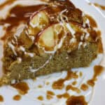 Close-up image of a slice of rustic apple and cornmeal cake drizzled with cider caramel and crème fraîche on a white plate.