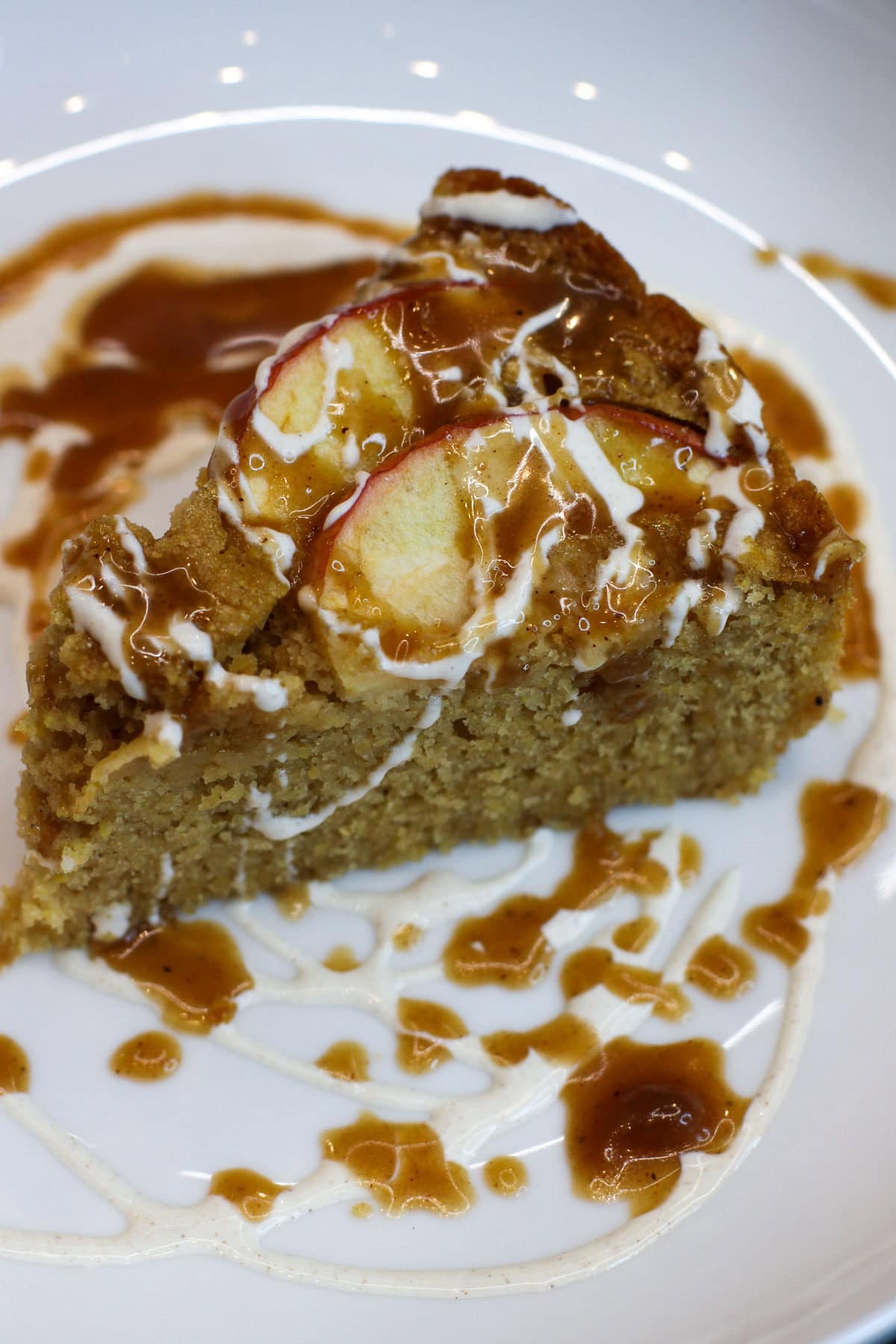 Close-up image of a slice of rustic apple and cornmeal cake drizzled with cider caramel and crème fraîche on a white plate.