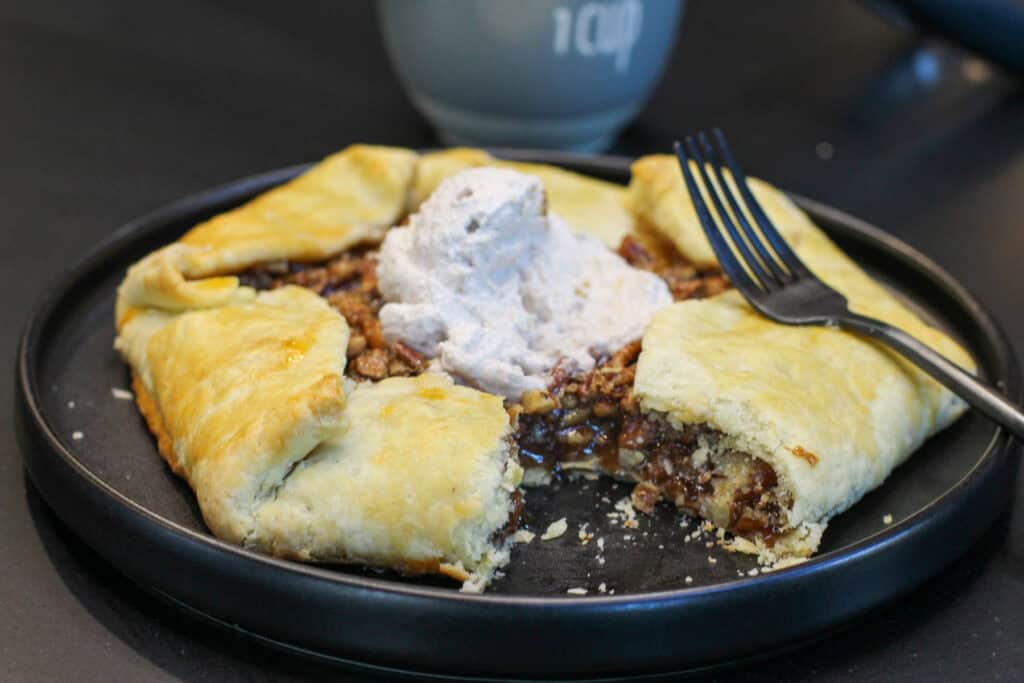 A golden-brown salted maple pecan galette with glossy caramelized filling and flaky folded crust, served with cinnamon whipped cream.