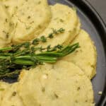 Round slices of savory herb shortbread made with rosemary, thyme, cheddar, and Parmesan arranged on a dark plate with fresh herbs.