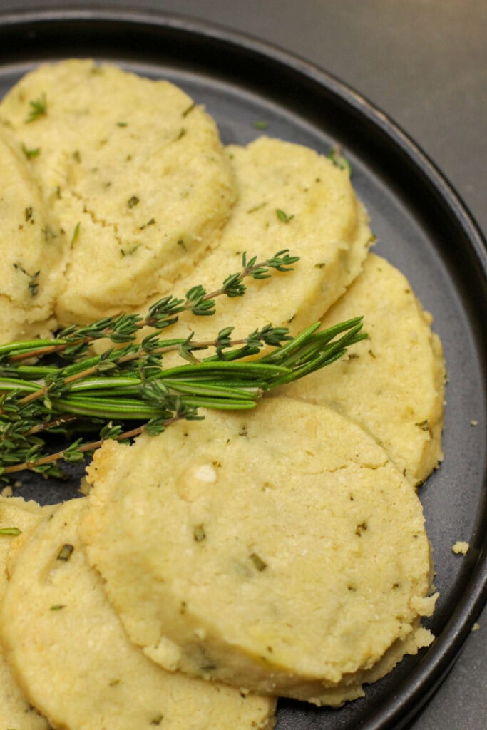 Round slices of savory herb shortbread made with rosemary, thyme, cheddar, and Parmesan arranged on a dark plate with fresh herbs.