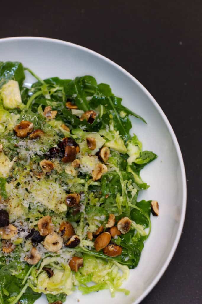 A fresh Brussels sprouts salad topped with shaved Pecorino Romano, toasted hazelnuts, and lemon-garlic dressing, served in a white bowl.