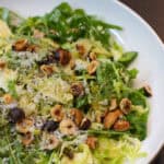 A fresh Brussels sprouts salad topped with shaved Pecorino Romano, toasted hazelnuts, and lemon-garlic dressing, served in a white bowl.