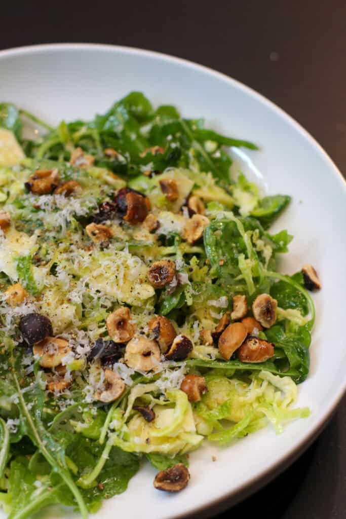 A fresh Brussels sprouts salad topped with shaved Pecorino Romano, toasted hazelnuts, and lemon-garlic dressing, served in a white bowl.