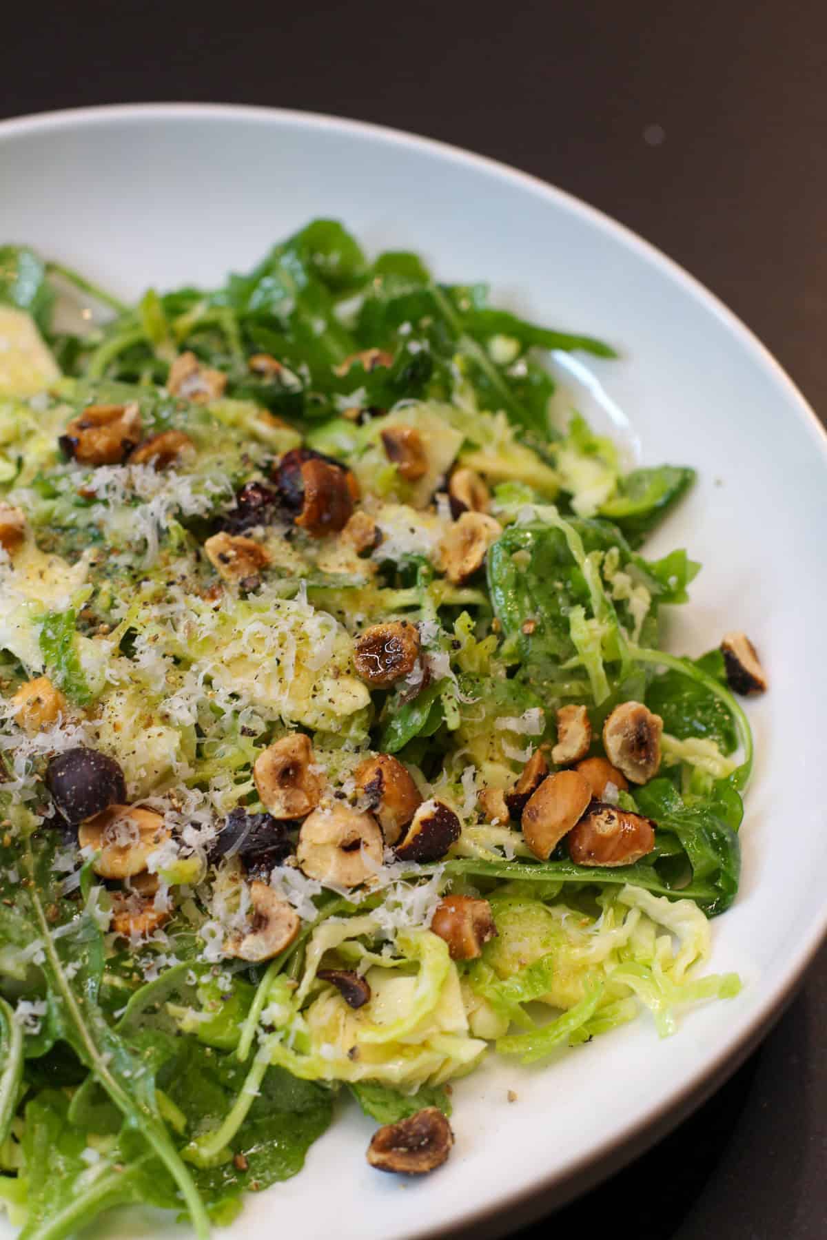 A fresh Brussels sprouts salad topped with shaved Pecorino Romano, toasted hazelnuts, and lemon-garlic dressing, served in a white bowl.