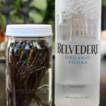 A glass jar filled with vanilla beans submerged in vodka, resting beside a bottle of organic vodka, showing homemade vanilla extract in progress.
