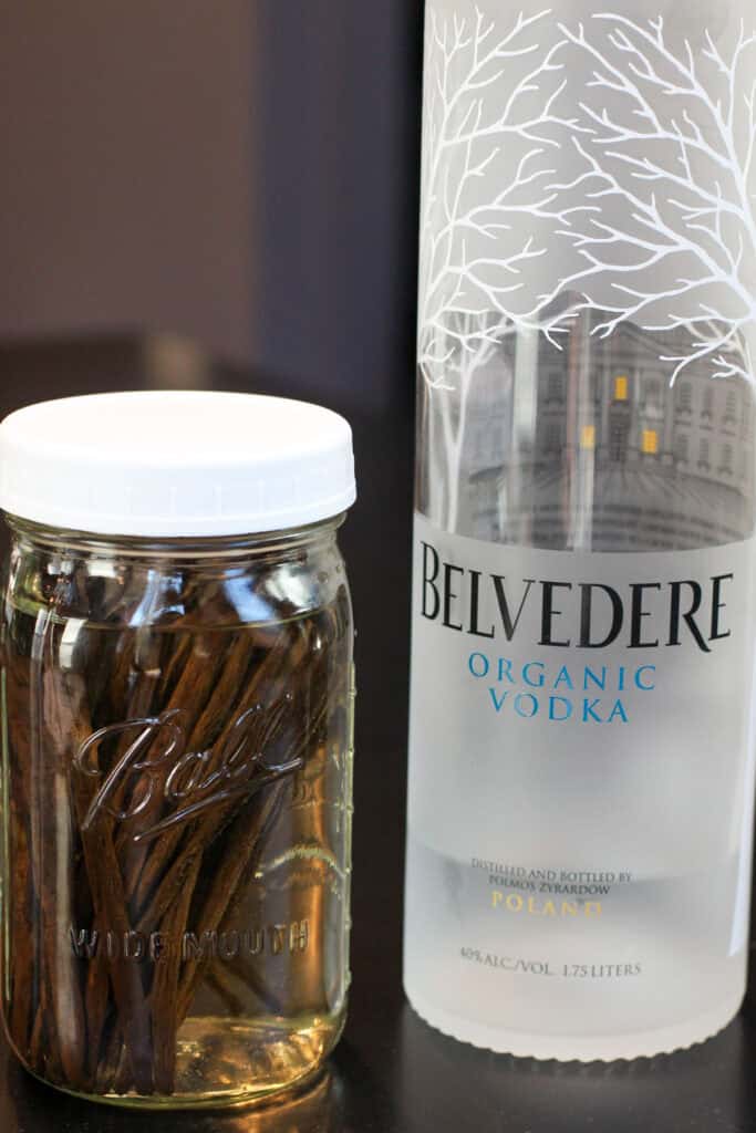 A glass jar filled with vanilla beans submerged in vodka, resting beside a bottle of organic vodka, showing homemade vanilla extract in progress.