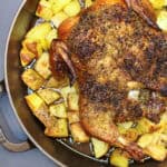 Golden whole roasted chicken seasoned with herbs, resting over crispy olive-oil–tossed potatoes in a cast-iron skillet. The chicken has crisp skin and juicy drippings surrounding the potatoes, creating a rustic, cozy dinner-party presentation.