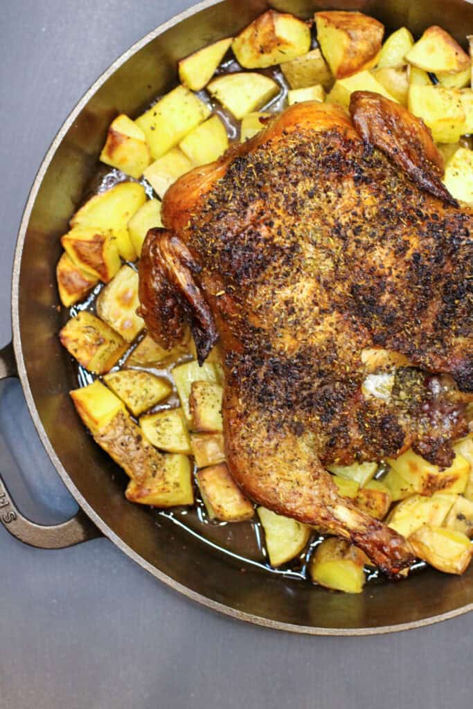 Golden whole roasted chicken seasoned with herbs, resting over crispy olive-oil–tossed potatoes in a cast-iron skillet. The chicken has crisp skin and juicy drippings surrounding the potatoes, creating a rustic, cozy dinner-party presentation.