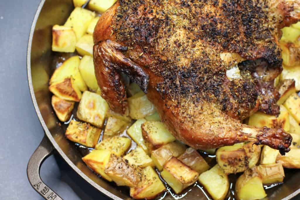 Golden whole roasted chicken seasoned with herbs, resting over crispy olive-oil–tossed potatoes in a cast-iron skillet. The chicken has crisp skin and juicy drippings surrounding the potatoes, creating a rustic, cozy dinner-party presentation.