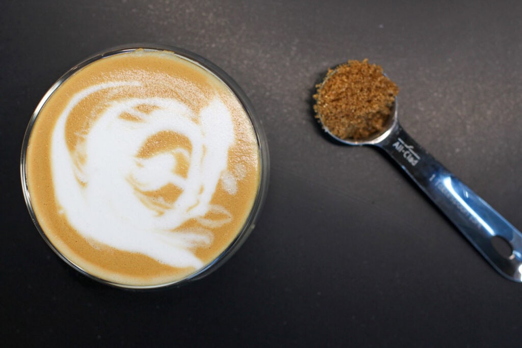 Brown sugar latte made at home with espresso and frothed milk in a glass cup on a dark background next to a tablespoon of brown sugar in a measuring spoon