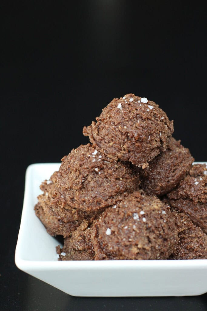 Chocolate sea salt protein bites stacked in a white serving bowl, made with almond flour and chocolate protein powder, finished with a light sprinkle of flaky sea salt on a dark background.
